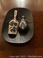 Two enamel decorated metal items placed on a black carved tray: a miniature scoop and a miniature flask