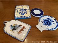 Four assorted serving boards or wall mounts displayed on a wooden surface, showing various decorative hand-painted designs including birds and floral patterns.
