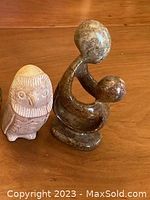 Two small stone sculptures displayed side by side on a wooden surface: a mother and child piece in dark brown polished stone and a hollowed owl figurine in lighter stone.