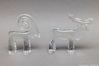 Two clear glass animal paperweights side by side, including a ram with spiral curved horns and an elk with large antlers, viewed from the front.