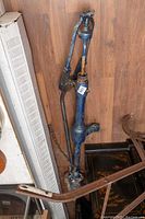 Full view of the tall blue cast iron antique well water pump leaning against a wooden wall, showing handle, cylinder, spout, and base.