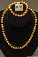 Full view of the pearl necklace arranged in a circle with the earrings centered on a black display, showing the golden color and uniform size of the pearls.