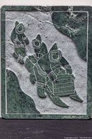 Full view of the verdi marble plaque showing three Inuit figures on sleds in carved relief with contrasting green and gray marble tones.