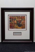 Framed artwork showing autumn landscape with colorful trees and a pool of water, bordered with matting and wooden frame.