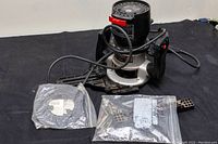 Full view of router with cord, circular guide ring wrapped in plastic, and plastic bag with multiple router bits.