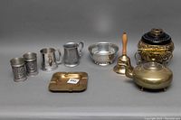 Photo showing entire lot on a grey background with brass, pewter, and silver items including teapot, bell, cups, bowl, and ashtray.
