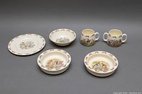 Five vintage porcelain Bunnykin nursery dishes: 2 feeding bowls, nursery plate, 2 two-handled mugs with Bunnykin illustrations.