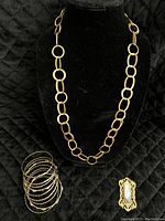 Full view of the vintage gold-tone open loop circle chain necklace, nine gold-tone bracelets stacked, and vintage rectangular brooch on black fabric background.