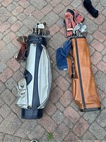 Two older golf bags with golf clubs outdoors on brick pavement.