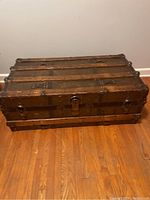 Front angled view of antique wooden shipping trunk on hardwood floor, showing wood slats and metal reinforcements with locks and latch.