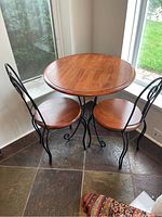 Full view of round wooden table with two wrought iron and wood chairs arranged around it inside near window.