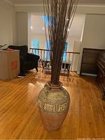 Full view of large ceramic vase filled with tall decorative sticks on hardwood floor.
