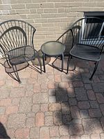 Two black metal mesh outdoor patio chairs with fan-back design and a small round metal table with glass top, positioned on outdoor stone patio.