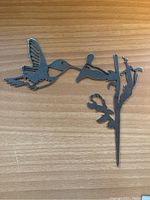 Metal hummingbird feeding from a flower on a stake, shown on a wooden surface to display details and size.