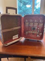 Open mini briefcase bar showing six glasses secured by beige elastic straps and metal bar accessories inside pink quilted lining.