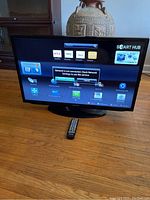 Front view of the smart TV powered on showing Smart Hub interface with apps like Netflix and Amazon Prime Video, remote placed in front on wooden floor.