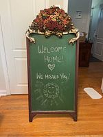 Frontal view of the decorative sandwich board chalkboard with floral carved top and green chalkboard surface showing chalk writing.