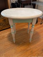 Front angle view of round solid wood end table painted white with mint green accents. Shows scratches and wear on top surface and legs.