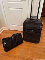 Black wheeled suitcase with retractable handle upright on wooden floor next to black carry bag.