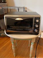Front view of Black & Decker toaster oven on a round table showing glass door, control knobs, and overall shape.
