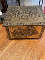 Firewood box showing the front panel with countryside cottage embossing and brass clad detail on caster base.