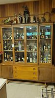 Front view of two-piece wooden china cabinet with glass door upper section displaying decorative bottles and items. Solid door bottom section with drawers in center.