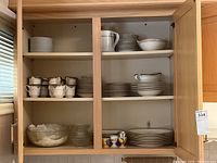 Open cabinet showing multiple shelves with Noritake Mi Amor cups, saucers, plates, Empress Bridal Gold plates, and a few glass bowls on lower shelf.