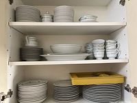 Shelf with three levels showing various stacks of plates, bowls, cups, and a yellow plastic tray with flatware inside.