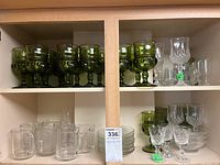 Shelf with multiple green embossed glass goblets, clear crystal-style goblets, clear mugs, and small clear plates.