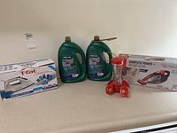Photo showing the T-Fal iron box, two green Kirkland dishwasher gel bottles with pumps, red hummingbird feeder, and Dirt Devil handheld vacuum in box arranged on a countertop.
