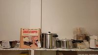 Photo showing Rival Grind-O-Matic grinder/chopper in box, pressure cooker, stock pot, and additional kitchen appliances on a shelf.