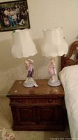 Pair of lamps on wooden nightstand, featuring boy and girl porcelain figurine bases with white fabric lampshades.