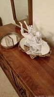 Photo showing porcelain figurine atop a rectangular mirrored vanity tray with gold-tone lipstick holder on wooden dresser
