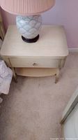 Full view of a beige night stand with a lamp on top, showing the drawer front and lower shelf.
