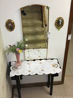 Full view of the table topped with doily, pink glass vase with faux florals, gold tone framed wall mirror hanging above, and two small oval gold framed wall art pieces on either side of mirror.
