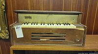 Front view of Emenee table top organ showing wooden body, keyboard with white and black keys, speaker grille with mesh and brass Emenee nameplate, visible damage including hole and white mold.