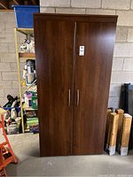 Front view of closed MDF wood-finished cabinet with two doors, silver metal handles, stands on a concrete floor against a block wall.