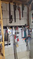 Overview of four pipe wrenches hanging on the wall and several clamps beneath them including bar clamps and quick grip clamps.