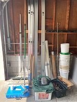 Photo showing a collection of metal wall studs, buckets, various long-handled tools, blue toolbox, plastic basket, and tarping rolls, arranged against a wooden wall.