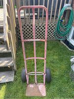Full front view of the metal dolly standing upright on grass showing pink tubular frame and two black wheels.