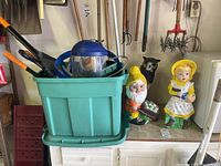 Green plastic tote filled with various garden tools including hand seed spreader, loppers, face protector, and others, alongside three garden figures: girl, gnome, and owl, arranged on a concrete surface in garage.