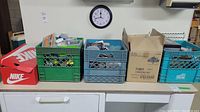Photo showing multiple containers including a red Nike box, green and blue crates, and a cardboard box on a white cabinet, with a clock on the wall.