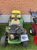 Front view of John Deere green ride-on mower with yellow seat, black steering wheel, Briggs & Stratton engine visible, manual on ground.