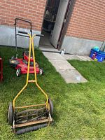 Photo showing both the yellow Turfglider push reel mower with visible rust and the red gas-powered Mastercraft lawn mower outside near a building.