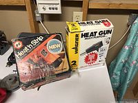 Two boxed products: Black & Decker Heat'n Strip electric paint remover and Wagner Heat Gun, on a white table surface.