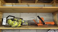 Yellow gas-powered chainsaw with black handle and worn yellow body showing scratches and rust, next to orange hedge trimmer with black handle and 16 inch blade