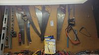 Photo of various hand saws and hand levels hanging on a pegboard wall, showing a mix of wooden and plastic handles, including large and small saws and several levels in metal and plastic.