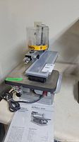 Front and side angled view of Ryobi 16-inch Scroll Saw SC160 showing motor, blade guard, blade clamp, tabletop, power cord and user manual on work surface.