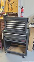 Photo showing two grey metal tool boxes stacked, one with multiple drawers, the other larger with a door and single drawer, on wheels in a workshop setting.