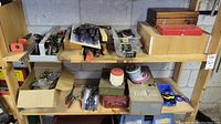 Wide view of two wooden shelves displaying various hand tools, small hardware boxes, and plastic bins containing nails, screws, and miscellaneous items. Tools include files, pliers, scissors, clamps, and tape measures.